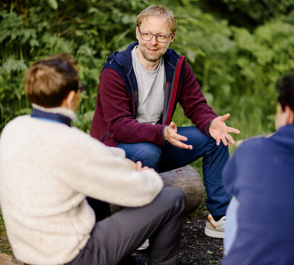Mann im Gespräch mit zwei Personen im Freien, umgeben von grüner Vegetation.