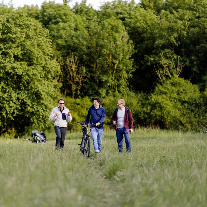 Waldspaziergang Drei Personen gehen auf einem Weg durch eine grüne Wiese, eine Person hat ein Fahrrad dabei.