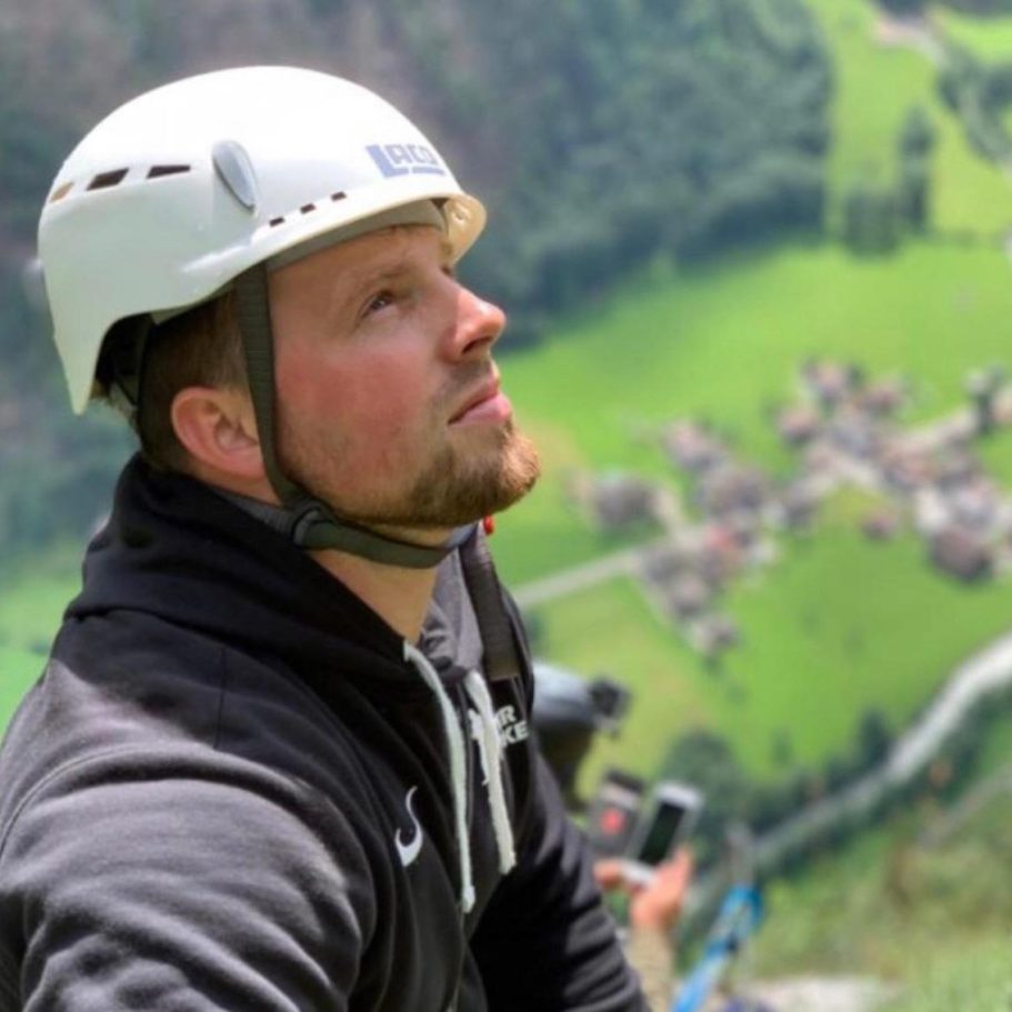 Mann mit Brille und Pullover, lächelt vor einer grünen Hintergrundlandschaft.