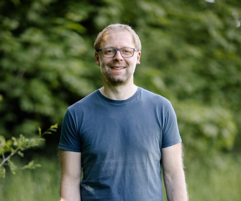 Coach Lächelnder Mann mit Brille, stehend vor grüner, bewaldeter Kulisse.