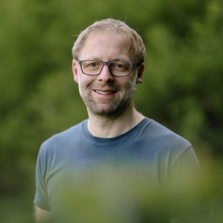 Männlicher Porträt mit Brille, lächelnd vor unscharfem grünem Hintergrund.